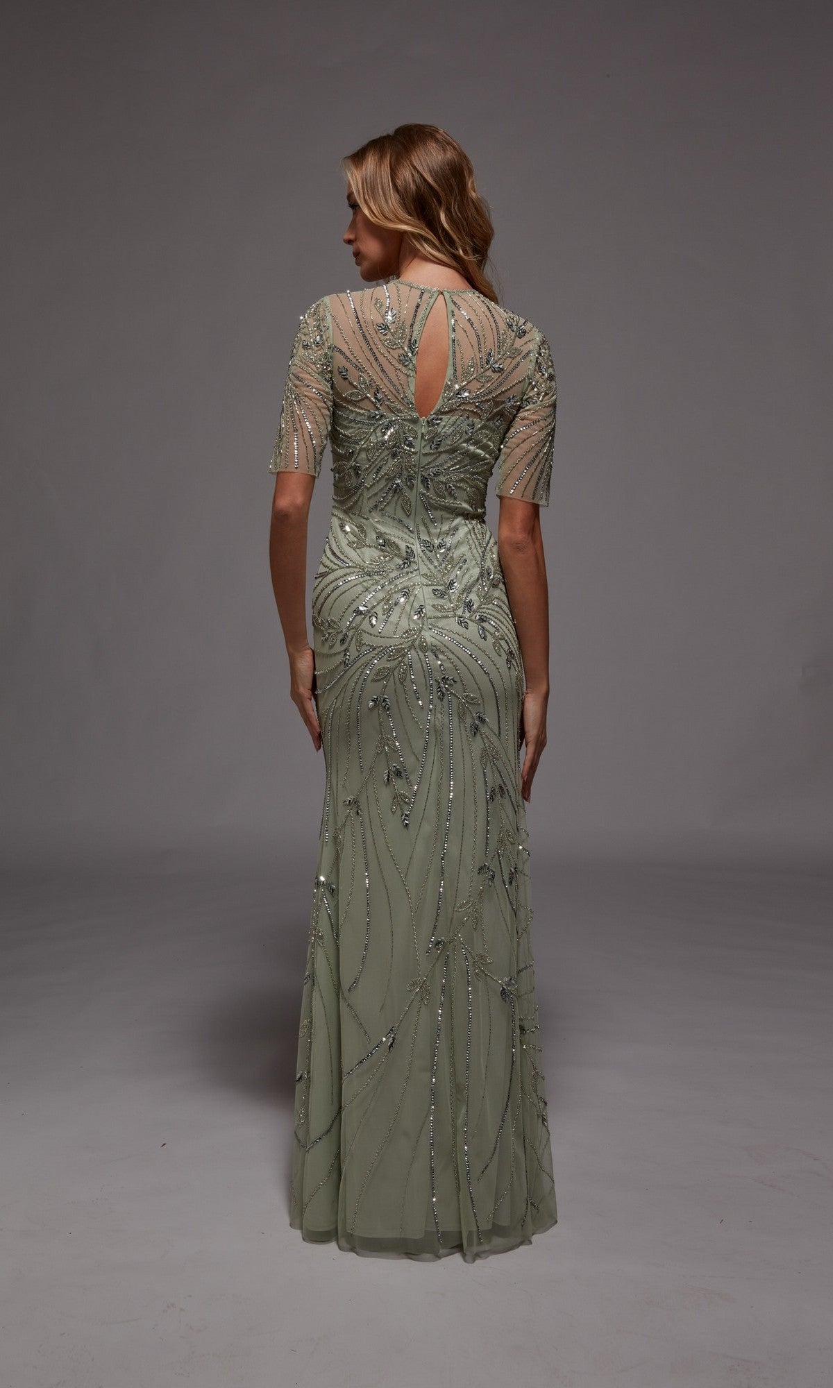 A woman faces away, wearing the Alyce 27809 long formal dress in light sage green, featuring sheer short sleeves and intricate beadwork—ideal for formal occasions or as a stunning mother-of-the-bride gown.